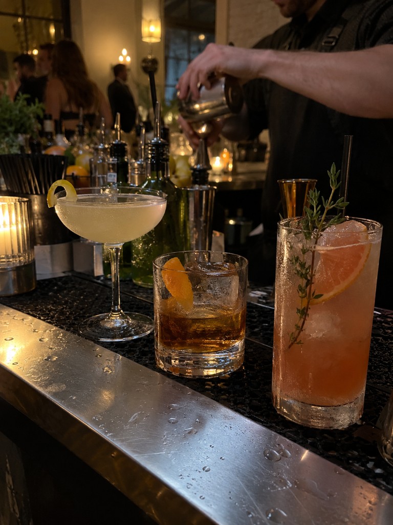 Coupe, old fashioned, and highball lined on a bar while a bartender pours